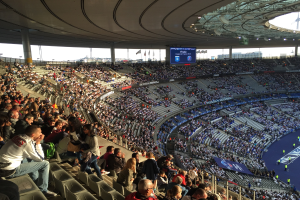 Große Menschenmenge in einem Stadion bei einem Fußballspiel mit einer Bühne, Flaggen, Stangen, einem Bildschirm und der Allianz Arena in München, Deutschland im Hintergrund.