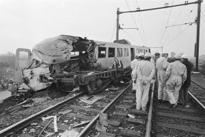 Eine Schwarz-Weiß-Szene eines Zugunglücks mit Menschen in Helmen drumherum, Strommäste, Bäume, Gebäude und einen klaren Himmel im Hintergrund.