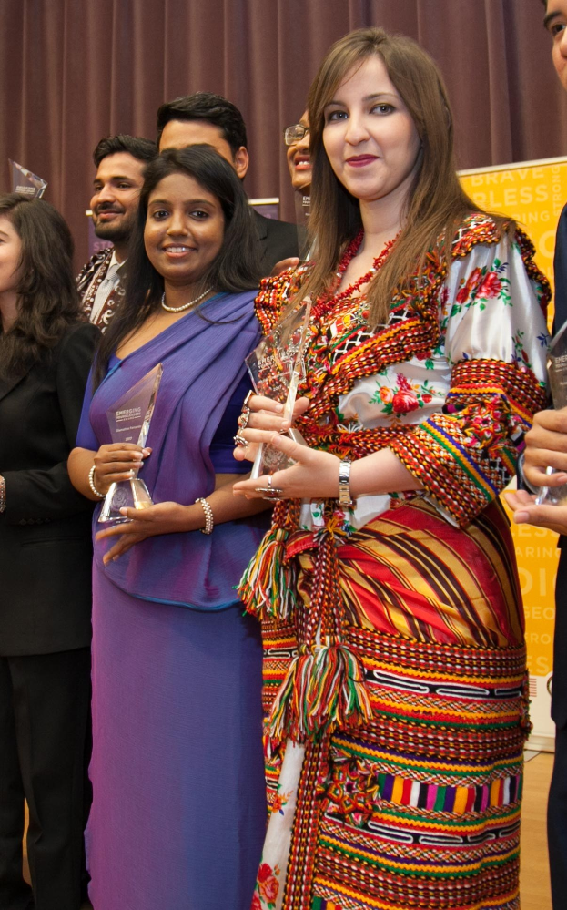 Gruppe von Menschen auf einer Bühne mit einer Frau in der Mitte, die eine Trophäe hält, vor einem Vorhang und einer Tafel mit der Aufschrift "Bravo Segenspreis-Gewinner."