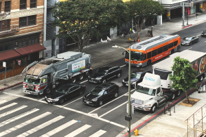Eine belebte Stadtstraße mit verschiedenen Fahrzeugen, Fußgängern, Gebäuden, Bäumen und Straßenmöblierung.