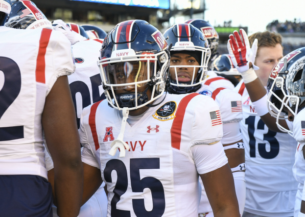 Gruppe von Männern in Football-Uniformen auf einem Feld mit Helmen und Handschuhen, Hintergrund zeigt unscharfe Zuschauer, ein Scoreboard mit 'Auburn vs Virginia Tech - 11/28/15 College Football Pick, Odds, and Prediction' und Himmel.