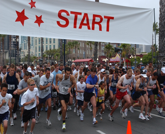 Gruppe von Läufern bei einem Marathon mit einem Verkehrskegel im Vordergrund und einem Banner mit Text im Hintergrund, umgeben von Bäumen, Laternenmasten, Gebäuden und einem klaren blauen Himmel.