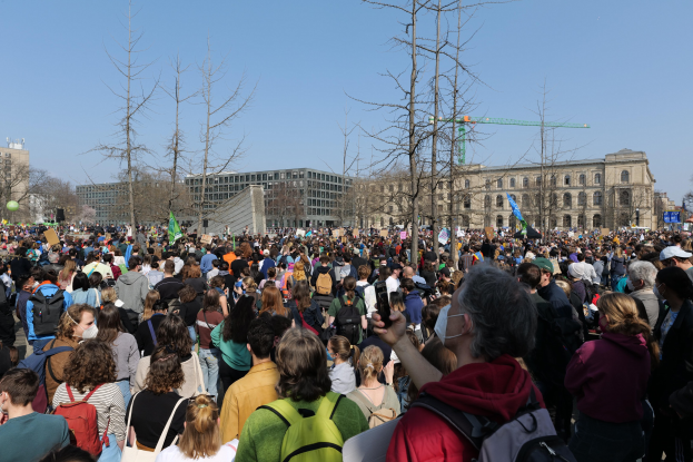Eine große Menschenmenge steht vor einem Gebäude mit Fenstern, Bäumen und einem klaren Himmel, viele halten Schilder und tragen Taschen, was auf eine Klimaprotest Demonstration hinweist.
