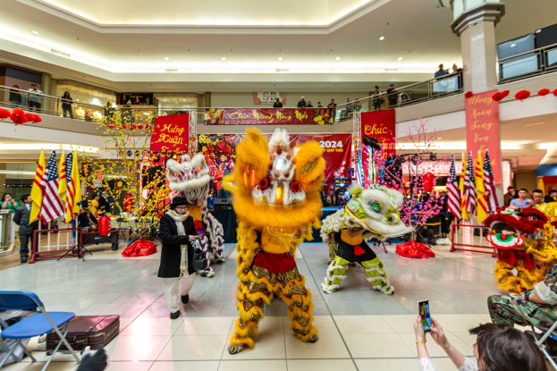 Vibrierendes chinesisches Neujahrsfest in einem Einkaufszentrum mit tanzenden Menschen in Kostümen und anderen, die auf Stühlen sitzen und Handys halten, umgeben von Fahnen, Bannern, Dekorationen, Geländern, Säulen und Deckenleuchten, mit einem Wasserzeichen unten.