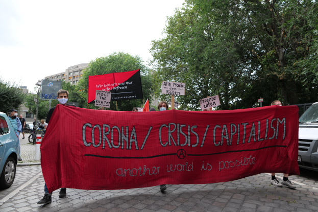 Eine Gruppe von Menschen marschiert mit einer roten Fahne, auf der 'Corona-Krise/Kapitalismus' steht, eine Straße entlang, mit parkenden Fahrzeugen, Gebäuden und Bäumen im Hintergrund unter einem klaren blauen Himmel.