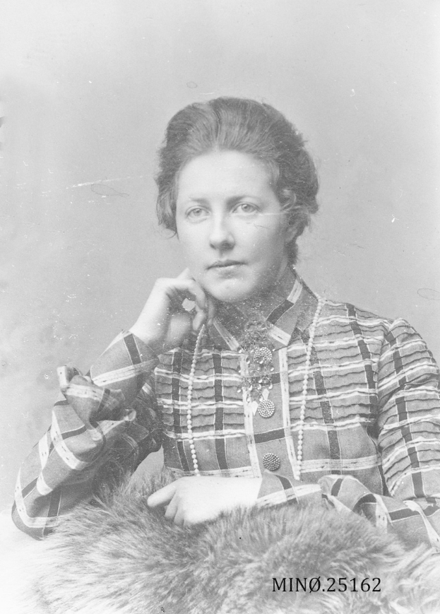 Ein Schwarz-Weiß-Foto einer Frau in einer karierten Bluse und einem Pelzcape mit zurückgebundenem Haar und einem ernsten und nachdenklichen Ausdruck.