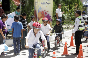 Kinder fahren auf Fahrrädern eine Straße mit Verkehrshütchen entlang, einige tragen Helme, andere stehen daneben, mit einer Fahne, Bäumen und Gebäuden im Hintergrund.