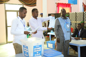 Eine Gruppe von Männern um einen Tisch mit verschiedenen Gegenständen, die bei einer Wahlurne ihre Stimmen abgeben, mit Menschen und Möbeln im Hintergrund.