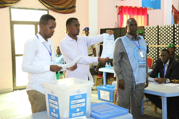 Eine Gruppe von Männern um einen Tisch mit verschiedenen Gegenständen, die bei einer Wahlurne ihre Stimmen abgeben, mit Menschen und Möbeln im Hintergrund.