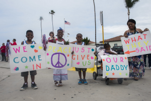 Gruppe von Kindern mit Schildern, auf denen "Wir kommen in Frieden, gebt uns unseren Daddy" steht, vor einem Gebäude, mit einer Frau, die einen Kinderwagen schiebt, umgeben von Bäumen, Pfählen, einer Flagge, einem Haus, Pflanzen und einem bewölkten Himmel.