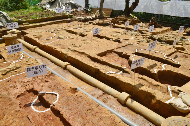 Ein großer Aushub mit Erde, Bäumen, Pflanzen, Gras, einem Zaun, einer Abdeckung, einer Wand, Tafeln mit Text, Rohren und anderen verstreuten Gegenständen.