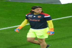 Ein Mann in einem blauen Hemd und gelben Shorts läuft auf einem Fußballfeld, die Arme ausgestreckt, mit entschlossener Miene und einem Tuch oben auf dem Bild.