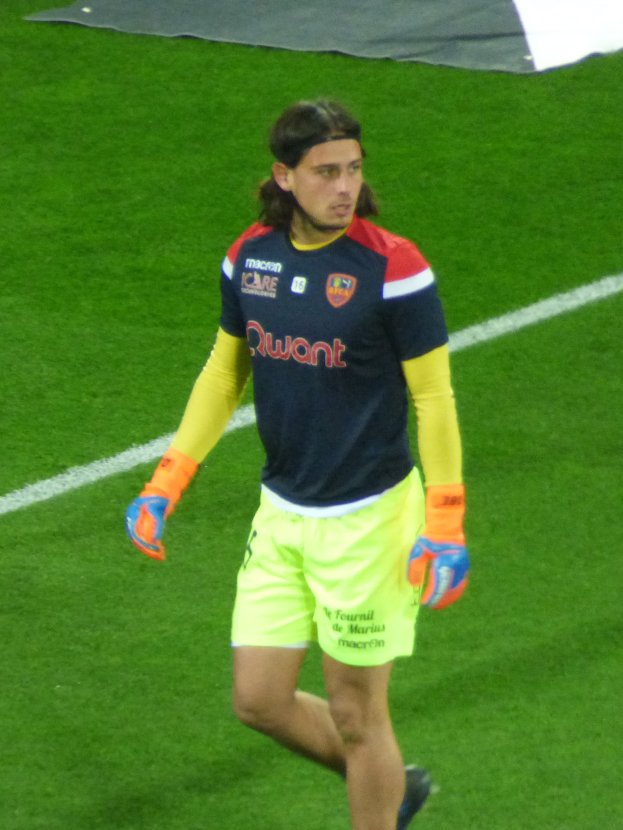 Ein Mann in einem blauen Hemd und gelben Shorts läuft auf einem Fußballfeld, die Arme ausgestreckt, mit entschlossener Miene und einem Tuch oben auf dem Bild.