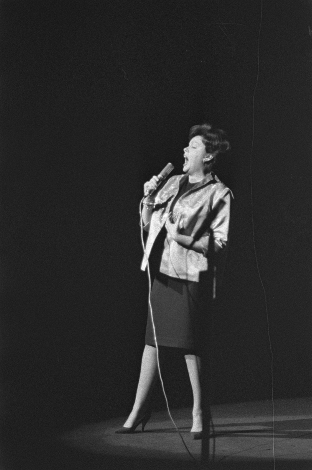 Eine Frau in einem schwarzen und weißen Outfit aus den 1950er-Jahren singt fröhlich auf einer von einem Scheinwerfer beleuchteten Bühne in ein Mikrofon, ihr Haar ist im klassischen 1950er-Stil frisiert.