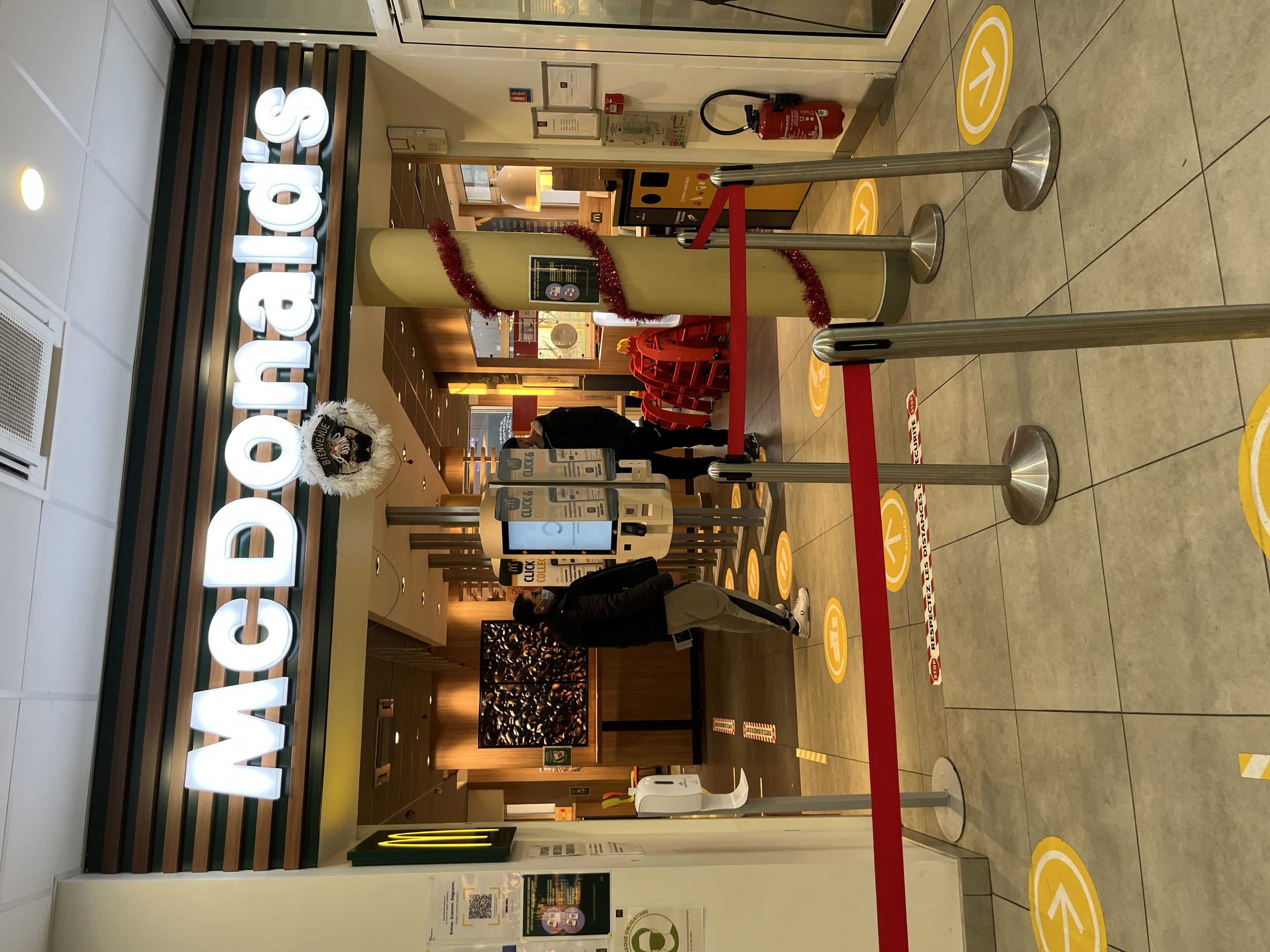 Innenseite eines McDonald's-Restaurants mit einem sichtbaren Schild, Metallstangen mit roten Bändern und einigen Personen im Vordergrund.