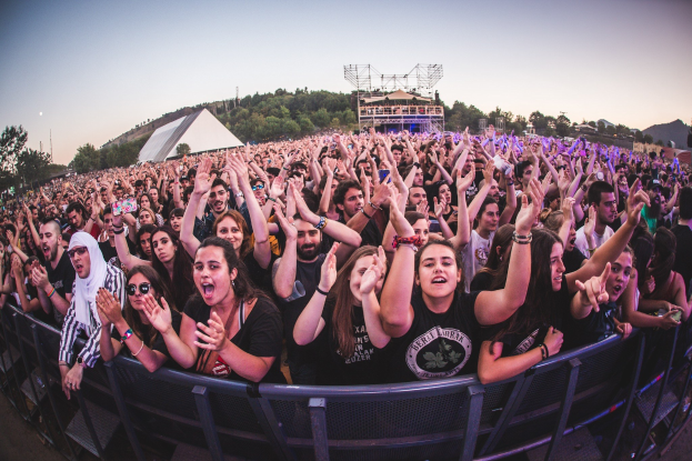 Eine gro├če Menge auf einem Musikfestival mit erhobenen H├Ąnden vor einem Gel├Ąnder, B├Ąume und Geb├Ąude sind im Hintergrund unter einem klaren blauen Himmel zu sehen.