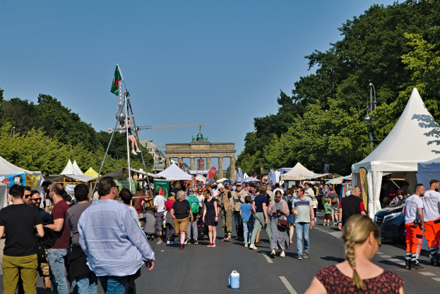 Eine Menschenmenge, die eine Straße entlanggeht, gesäumt von Zelten, Fahrzeugen und Bäumen, mit einem Bogen und einem klaren blauen Himmel im Hintergrund und Fahnenmasten auf der linken Seite.