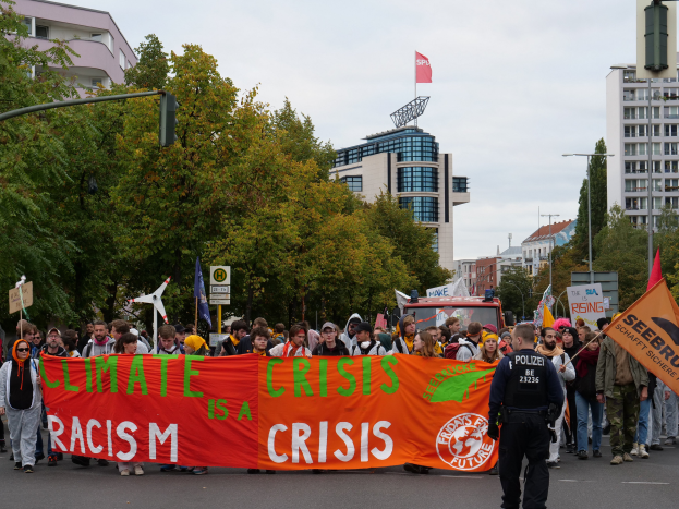 Eine Gruppe von Menschen marschiert eine baumbestandene Straße entlang und hält ein Banner mit der Aufschrift "Klimakrise ist eine Krise", mit Gebäuden und einem klaren blauen Himmel im Hintergrund.