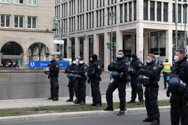 Polizeibeamte in schwarzen Uniformen und Masken stehen vor einem Berliner Gebäude mit Glasfenstern und Säulen, einige halten Helme, mit Laternen, Verkehrszeichen, Schildern mit Text, einer Statue und anderen Menschen im Hintergrund.