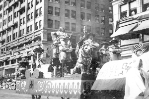 Schwarzes und weißes Foto eines Umzugswagens, der mit Statuen von Menschen und Tieren geschmückt ist und die Straße einer Stadt entlangfährt, während Zuschauer von der Straße aus zusehen.
