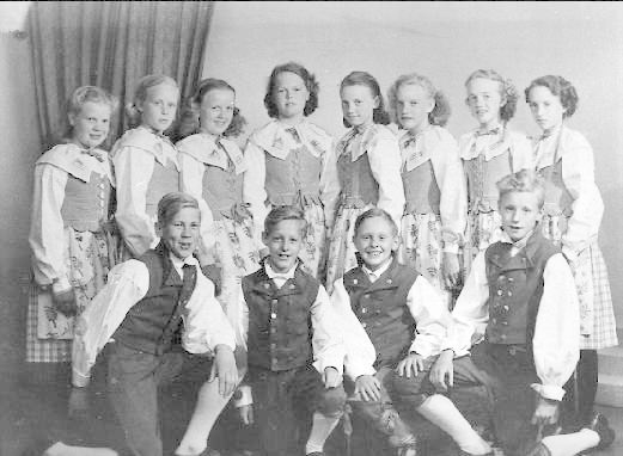 Schwarz-weiß-Foto einer Gruppe von Kindern, die vor einer Wand mit einem Vorhang posieren, mit dem Text "1930er-Kinder-Schule" unten.