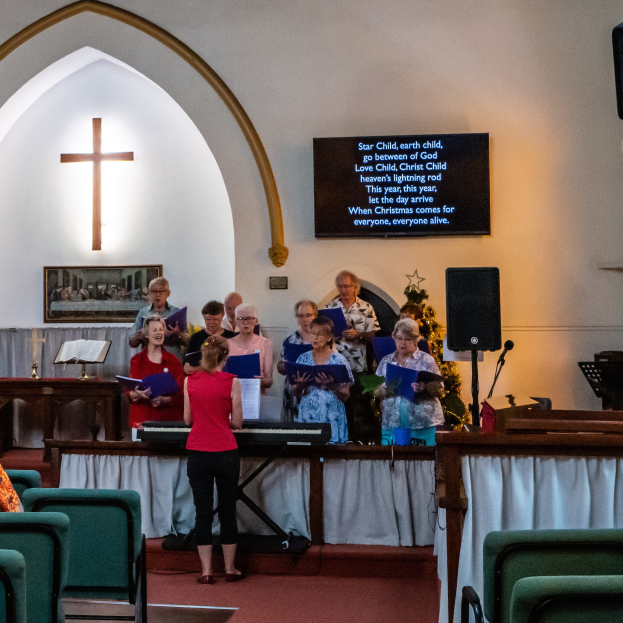 Eine Gruppe von Menschen steht vor einer Kirche, einige halten Bücher, Stühle links, ein Kreuzsymbol und ein Photo-Rahmen an der Wand, eine Tafel mit Text, ein Lautsprecher mit Ständer und ein Tisch mit einer Decke, scheinbar singend als Chor.