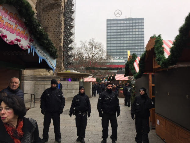 Polizisten in schwarzen Uniformen stehen vor einem geschmückten Weihnachtsmarkt in Berlin mit festlichen Buden, Gebäuden, Bäumen und einem klaren Himmel im Hintergrund.