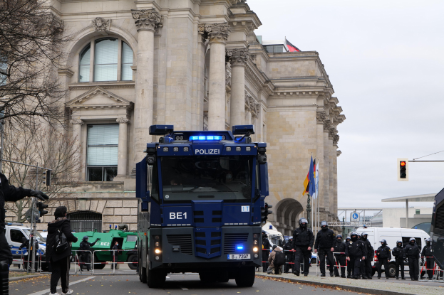 Eine Gruppe von Polizisten steht vor einem großen Gebäude mit Fenstern, Pfeilern und Bogengängen, mit Fahrzeugen auf der Straße und einer Person mit einer Kamera links; Trees, Verkehrssignale, Fahnenmäste und ein klarer blauer Himmel sind im Hintergrund zu sehen.