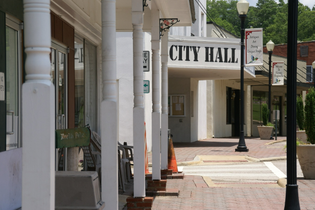 Rathaus mit umliegenden Gebäuden, Straßeninfrastruktur, Kraftfahrzeugen, Absperrgittern, Möbeln, Grünpflanzen und einem bewölkten Himmel.