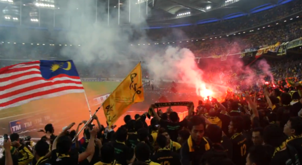 Eine große Menge Menschen in einem Stadion mit Bengalos, Fahnen und einer Kamera, mit Rauch und Schildern mit der Aufschrift "Malaysia vs. Malaysia Live-Streaming" im Hintergrund.