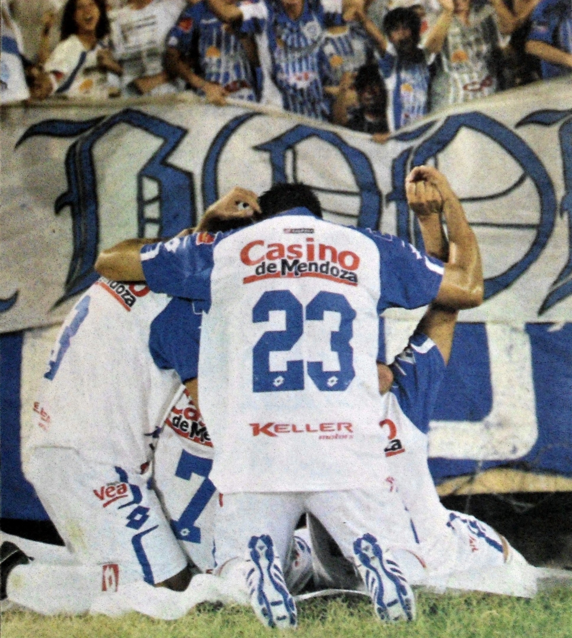 Gruppe von Fußballspielern, die vor jubelnden Zuschauern auf dem Boden feiern, mit einem Banner mit Text im Hintergrund.