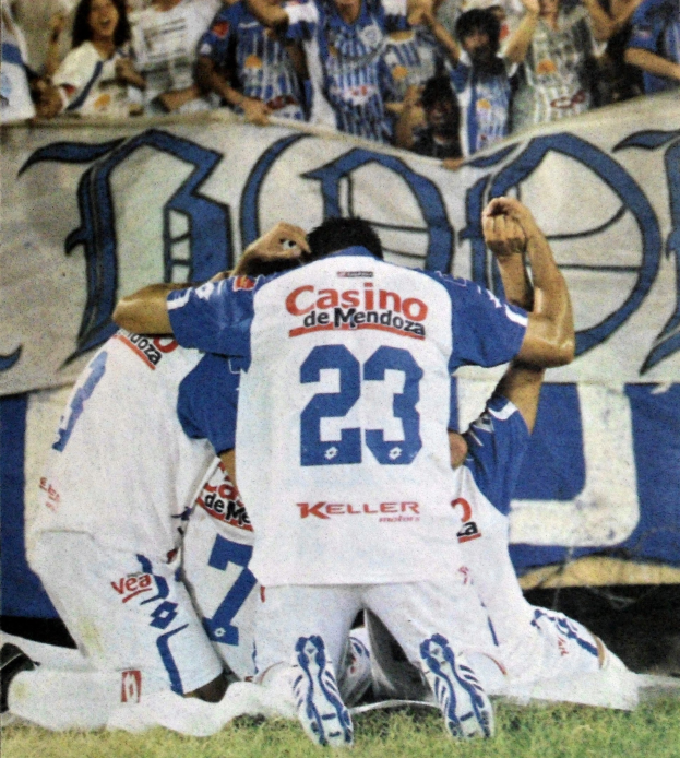 Gruppe von Fußballspielern, die vor jubelnden Zuschauern auf dem Boden feiern, mit einem Banner mit Text im Hintergrund.