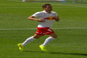Ein Mann in einem weißen Hemd und roten Shorts, der auf einem Fußballfeld läuft, mit einem Torpfosten im Hintergrund und ein paar Menschen in der Nähe, die Fußballschuhe tragen.