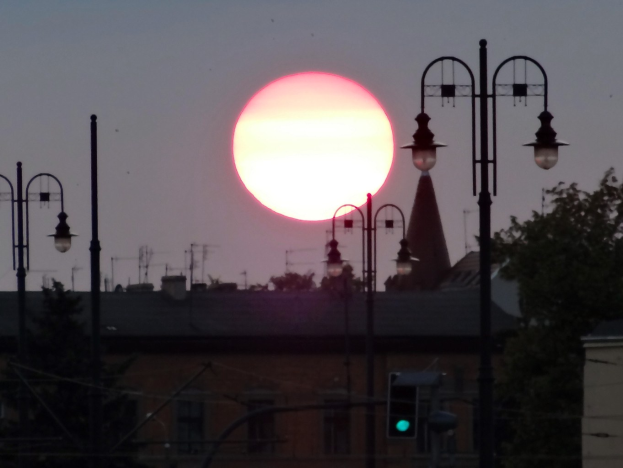 Eine Straße in einer Stadt bei Sonnenuntergang mit Straßenlaternen, einer Ampel, Bäumen, Gebäuden, Masten und Antennen im Hintergrund, die in warmem Orange leuchten.