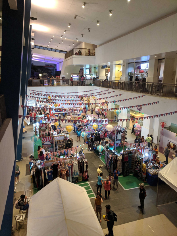 Eine große Menge bewegt sich durch einen belebten Einkaufsmarkt mit Kleiderständen, festlichen Dekorationen und Sitzbereichen, was auf einen Weihnachtsmarkt im Mall of Asia hindeutet.