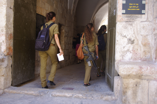 Israeli soldiers in uniform walk through an archway in Jerusalem's old city, one holding a gun and a paper, with a board on the wall to the right.