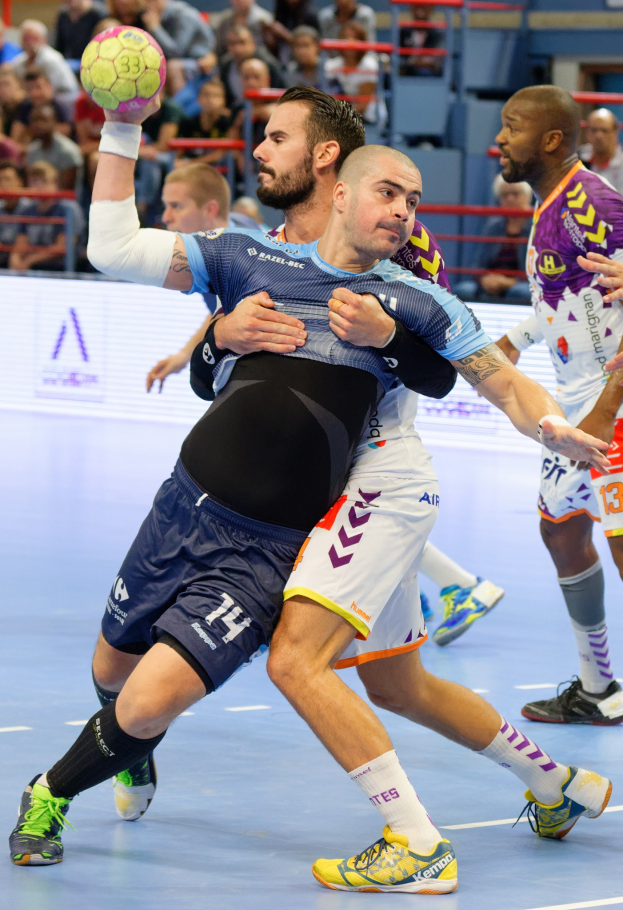 Gruppe von Männern, die Handball spielen, einer hält den Ball, mit Zuschauern im Hintergrund.