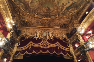 Das Innere eines Theaters mit einem roten Vorhang, Kronleuchtern, Skulpturen, Deckenmalerei und einer Bühne mit einigen Menschen davor.