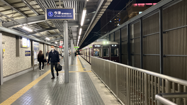 Ein Zug fährt auf einem Bahnsteig ein, mit Menschen, die gehen, einem Geländer auf der rechten Seite, Gebäuden im Hintergrund und Lampen mit einer Anzeigetafel oben auf dem Bahnsteig.