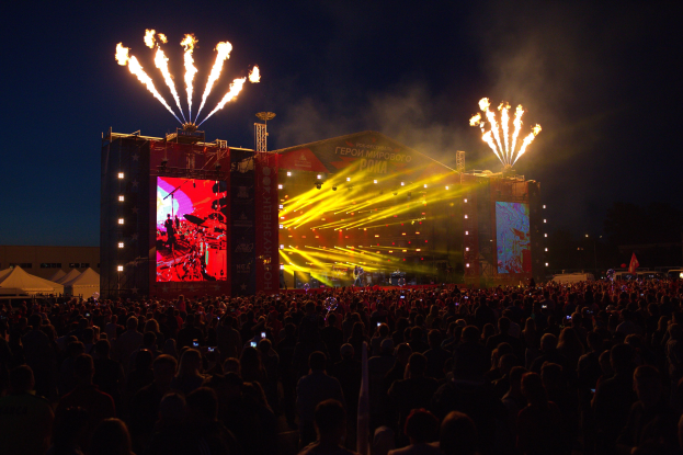 Eine große Menschenmenge bei einem Musikfestival schaut auf eine hell erleuchtete Bühne mit Bannern, Zelten links und Feuerwerk am dunklen Himmel.