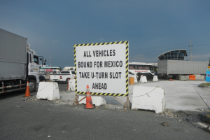 Lastwagen auf einem Parkplatz neben einem Schild mit der Aufschrift "Alle Fahrzeuge nach Mexiko" mit verstreuten Verkehrskegeln, Brettern und Hintergrundgebäuden, Bäumen, Polen und einem klaren blauen Himmel.