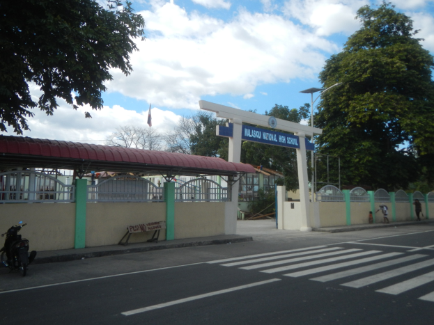 Einfahrt zu einer Schule mit einem vor der Tür geparkten Motorrad, mit einem Tor, einem Schild, Straßenmarkierungen, Laternen, Bäumen, einer Fahne und einem bewölkten Himmel.