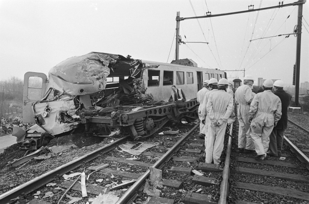 Eine Schwarz-Weiß-Szene eines Zugunglücks mit Menschen in Helmen drumherum, Strommäste, Bäume, Gebäude und einen klaren Himmel im Hintergrund.