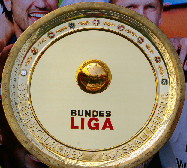 Ein Mann hält ein goldenes Schild mit 'Bundesliga' darauf, mit einem Banner mit drei Personen im Hintergrund.