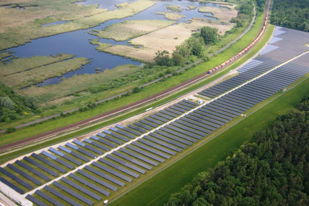 Eine Luftaufnahme eines Solarparks mit zahlreichen Solarmodulen auf einem Feld, umgeben von Bäumen, Gras, Wasser und einer nahen Bahntrasse.
