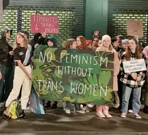 Eine Gruppe von Menschen mit einem Banner "Kein Feminismus ohne Transfrauen" und Schildern in einem öffentlichen Raum mit einer Wand und einem Rollladen im Hintergrund.