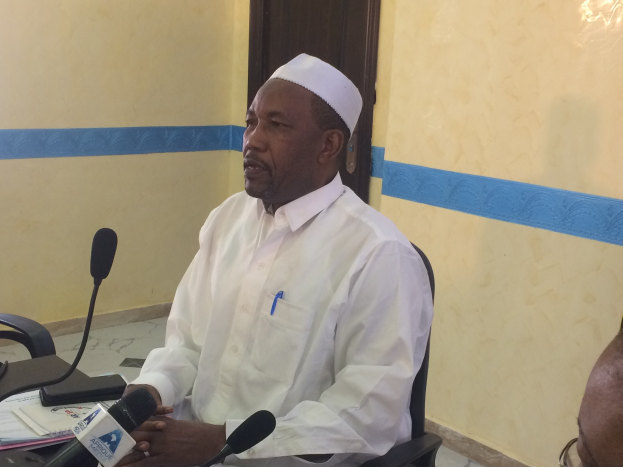 Ein Mann in einem weißen Kleid und Hut sitzt an einem Tisch mit Mikrofonen, spricht, während Papiere und Gegenstände auf dem Tisch zu sehen sind, mit einer anderen Person mit Brille rechts von ihm, in einem Raum mit einer Wand und einer Tür im Hintergrund.