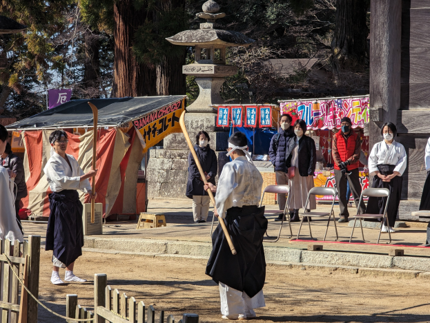 Eine Gruppe von Menschen in traditioneller Kleidung versammelt sich im Freien in Kyoto, einige tragen Masken und halten Holzstöcke, mit Stühlen, Bannern und einem Zelt im Hintergrund unter einem klaren blauen Himmel.