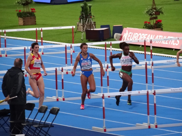 Eine Gruppe von Frauen, die Hürden auf einem Rasenlaufbahn läuft, mit einer Person, die links eine Stafettenstab hält, Stühle und ein Schreibtisch im Vordergrund, Blumentöpfe im Hintergrund und Schilder mit Text.