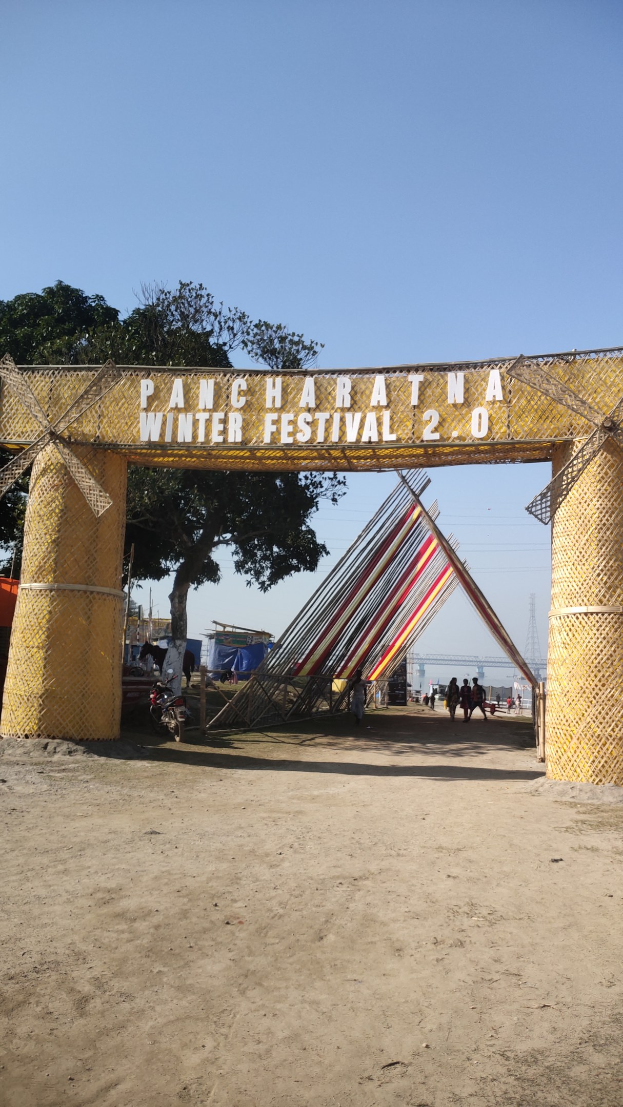 Eine festliche Szene mit einem Riesenrad in der Mitte, einem Tor mit Text, einer Gruppe von Menschen, Pfählen, einem Turm, Bäumen, einem Zaun und einem bewölkten Himmel.